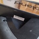 Detail černé plastové části hudebního příslušenství s kovovým elementem. Na kovovém povrchu je vidět nápis Roland a na bílé nálepce kód TY63498. Povr…