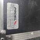 Detail kovového štítku na černém povrchu transportního kufru s hliníkovými hranami. Na štítku je uveden nápis PROEL, TECHNOLOGY AS ART a www.proel.co…
