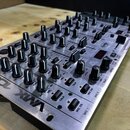 Stříbrný mixážní pult s kovovým povrchem a velkým množstvím černých otočných ovladačů. Na spodním okrave je nápis „VMA 1000“. Na panelu jsou viditeln…