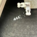 Detailní pohled na tmavý, texturovaný povrch. Na povrchu je bílým nápisem zapsáno číslo 444). V horní části je vidět část světlého plastového nebo ko…