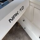 Detail bílého klavíru nebo syntezátoru s viditelnými černými a bílými klávesami. Na boku nástroje je černý nápis NPK-10. Povrch je hladký a v dobrém…