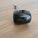 Černý plastový knoflík s nápisem Roland. Je připevněn na kovovou osa a položen na dřevěném povrchu. Povrch plastu vykazuje drobné škrábance.