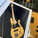 Fotografie zachycuje papírový tisk s vyobrazením elektrické basové kytary v žlutém provedení s viditnou strukturou dřeva. Nástroj má černý pickguard…