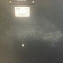 Detail zadní strany černého reproduktorového boxu s texturovaným povrchem. V horní části je kovový štitek s nápisem MADE IN ENGLAND a bílá nálepka s…