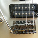 Detail elektrické kytary v stříbrné barvě. Je vidět černý humbucker snímač a chromovaný kobylník s páčkou. Na povrchu kytary jsou v okolí snímače vid…