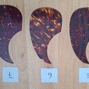 Tři kytarové plektra v tmavě hnědé a červené barvě s žlutými vzory (tortoise shell). Jsou umístěna vedle sebe na světlém dřevěném povrchu. Pod každým…