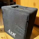Černý textilní kryt na hudební zesilovač nebo reproduktor. V dolní části je bílý nápis „AER“ a pod ním menší text „the acoustic people“. Horní strana…