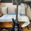 Černá elektrická kytara s ostrými hranami korpusu a špičatou hlavou. Na hrazence je vidět bílý nápis, který není čitelný. Kytara má dva humbuckery a…