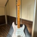 Elektrická kytara s bílým trupem a světlou dřevěnou hmatnicí. Na hlavě je viditelné logo Squier a nápis STRATOCASTER. Kytara má bílý pickguard, tři s…