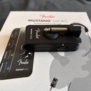 Černý kompaktní sluchátkový zesilovač s vloženým jack konektorem, položený na originální krabici. Na zařízení je bílý nápis Fender a MUSTANG MICRO. K…