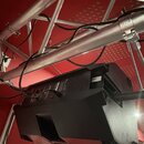 Černý aktivní reproduktor v plastovém korpusu, který je zavěšen pod hliníkovým rastrem. Na zadním panelu jsou viditelné konektory a štítky s technick…