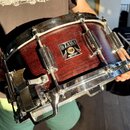 Červený dřevěný snare drum s kovovými obručemi a mechanismem na straně. Na korpusu je čitelný nápis TAMA SUPERSTAR a pod ním text MADE IN JAPAN. Povr…