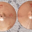 Dva mosazné činely typu hi-hat položené na šedém koberci. Levý činel nese nápisy „PAISTE 2002“, „TRADE MARK MADE IN SWITZERLAND“ a „TOP MEDIUM HI-HAT…