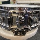 Stříbrný kovový snare drum s chromovaným povrchem. Na boku je viditelný oválný štítek s nápisy DYNA-SONIC, CUSTOM BUILT a nečitelným textem mezi nimi…