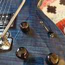 Detail elektrické kytary s modrým plamenovitým povrchem. Na snímačích je viditelný nápis Seymour Duncan. Kytara má tři černé ovládací knofíky a páčku…