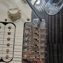 Detailní pohled na chromovaný kobylku elektrické kytary s páčkou. Na každém z šesti sedel je vyryt nápis FENDER. Kolem kobylky je vidět bílý pickguar…