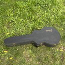 Černý tvrdý kufr na kytaru s bílým prošíváním po okrajích. Na horní části je vyražený nápis Gibson. Kufr leží na travnatém povrchu a má viditelné kov…