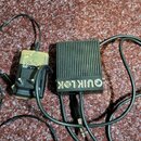 Černý kovový box s žebrovaným povrchem a nápisem QUIK LOK. K zařízení je připojen napájecí adaptér béžové barvy a černý kabel s jack konektorem. Povr…