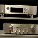 Dvě stříbrná audio zařízení značky marantz umístěná v policích. Horní přístroj má nápis NETWORK AUDIO PLAYER NA6005, displej a ovládací prvky. Dolní…
