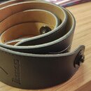 Černý kytarový popruh z kůže, srolovaný do spirály na dřevěném povrchu. Na popruhu je vyražené logo a nápis Ibanez. Konec popruhu je zakončen otvoru…