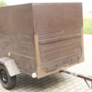 Hnědý přívěsný vozík s uzavřenou skříní. Spodní část stěn je tvořena vodorovnými dřevěnými prkny, horní část je z kovového plechu. Na vozíku nejsou v…