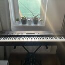Černý digitální klavír umístěný na černém X-stojanu. Na levé straně je viditelné logo YAMAHA a vpravo nápis PANORAMA. Instrument má bílé klávesy, ovl…