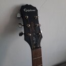 Černá hlava elektrické kytary s bílým logem Epiphone. Na povrchu jsou viditelné nápisy Junior MODEL a Les Paul JUNIOR. Lak na hlavě vykazuje několik…