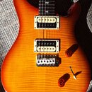 Elektrická kytara s dvojcutovým tvarem těla a povrchem v oranžovo-žlutém přechodu s viditnou strukturou dřeva. Nástroj má dva humbuckery s krémovými…