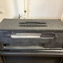 Černý kytarový kombozesilovač s texturovaným povrchem a kovovými rohy. Na horní straně je plastová rukojeť a ventilační mřížka. Zadní strana je otevř…