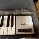 Černý klávesový nástroj s bílými a černými klávesami. Na horní liště je nápis String Performer. Ovládací panel obsahuje posuvník s označením VOLUME S…