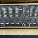 Šedý mixovací pult s velkým množstvím potenciometrů, posuvníků a vstupních konektorů. Na horní části je vidět bílý nápis ALLEN&HEATH a označení model…