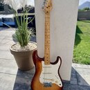 Elektrická kytara s trupem v barevnosti sunburst a hnedým hmatníkem. Na hlavě je viditelné logo Fender. Kytara má bílý pickguard, tři snímače a kovov…
