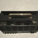 Černý kovový přístroj s ventilačními otvory na horní a boční straně. Na horní části je připevněna kovová rukojeť stříbrné barvy. Na zadní straně jsou…