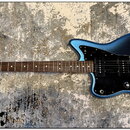 Modrá elektrická kytara s černým pickguardem a hnědým hmatníkem. Na hlavě je viditelné logo Fender a nápis Stratocaster. Kytara má jeden humbucker u…