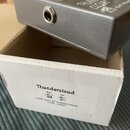 Stříbrný efekt pedál s kovovým povrchem umístěný na bílém kartonovém krabici. Na pedálu je viditelný nápis THUNDER CLOUD, KATANASOUND a OUTPUT u výst…