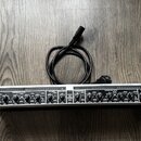 Stříbrný rackový procesor s černými ovládacími prvky a nápisem COMPOSER PRO-XL. Na pravé straně je logo behringer a vypínač POWER. Za přístrojem leží…