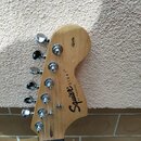 Záběr na hlavu elektrické kytary z přírodního dřeva. Na povrchu je černý nápis Squier® STRAT® a pod ním menší text BY FENDER®. V horní části je umíst…