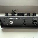 Zadní strana černého audio rozhraní s nápisem M-AUDIO. Na zadním panelu jsou umístěny USB konektor, dva výstupy MAIN L a R, vstup INPUT 1 s označením…