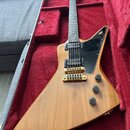 Elektrická kytara s výrazným asymetrickým tvarem těla v přírodním dřevě, položená v červeném plišovém kufru. Nástroj má dva humbuckery, zlatý most a…