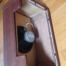 Fotografie zachycuje otevřený dřevěný reproduktorový box v hnědé barvě. Uvnitř je osazen černý reproduktor s bílo-modrým štítkem, na kterém je nápis…