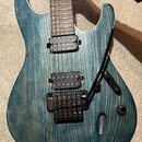 Elektrická kytara s modrozeleným povrchem s viditnou strukturou dřeva a bílým lemováním. Nástroj má dva černý humbuckery, černý most s pákou a jeden…