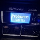 Detailní pohled na ovládací panel černého elektronického zařízení. Na horní části je bílý nápis PreSonus a AIR18s, který se opakuje i na podsvíceném…