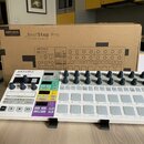 Bílý sekvencer Arturia BeatStep Pro umístěný na černém stojanu před svou originální kartonovou krabicí. Na přístroji jsou vidět šeradovky šedých pods…