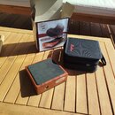 Na dřevěném stole je umístěn Stompbox v dřevěném provedení s černou horní plochou a nápisem "Shadow". Vedle něj je černý textilní přepravní pouzdro s…