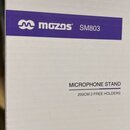 Bílá papírová krabice s fialovým pruhem. Na pruhu je bílé logo a nápis mgzos SM803. Níže je uveden text MICROPHONE STAND 200CM 2 FREE HOLDERS.