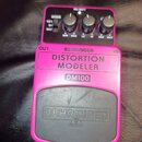 Růžový efekt pedál s nápisy BEHRINGER, DISTORTION MODELER a DM100. Na horní části jsou tři ovládací knoflíky s popisky MIN MAX, LO HI a MIN MAX, přep…