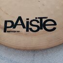 Detail kovového činely v zlatavé barvě. Na povrchu je černý nápis pAiste a pod ním menší text MADE IN GERMANY. V horní části loga jsou viditelné drob…