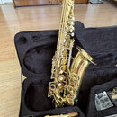 Zlatý saxofon v černém kufru s polstrováním. Na zvonu nástroje je vyryté logo a nápis "YAMAHA". V kufru vedle saxofonu leží munční dutina a část přís…