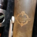Detailní záběr na dřevěný nástroj s hnědým povrchem. Na povrchu je zlatý nápis SUZUKI a pod ním menším písmem MUSIQUE, doplněný o dekorativní emblem…