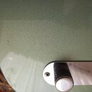 Detail zeleného lakovaného povrchu kytary s kovovým ovládacím panelem. Na panelu je vidět jeden černý vlněný knoflík a šroub. V laku je v levé části…